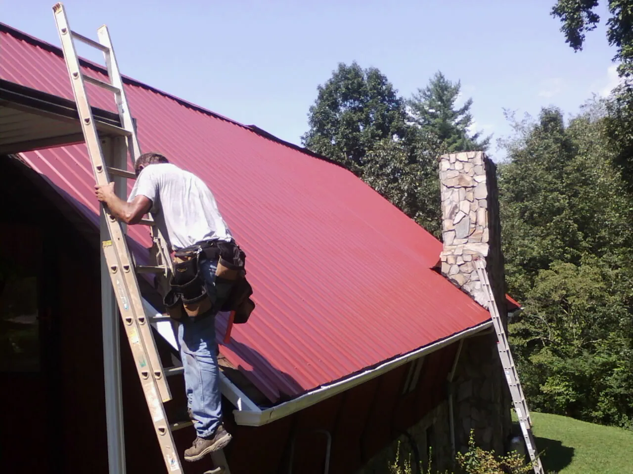 Roofer on a ladder beside a red metal roof and stone chimney — Soffit & Fascia Repair by Roofing Brothers in Patton